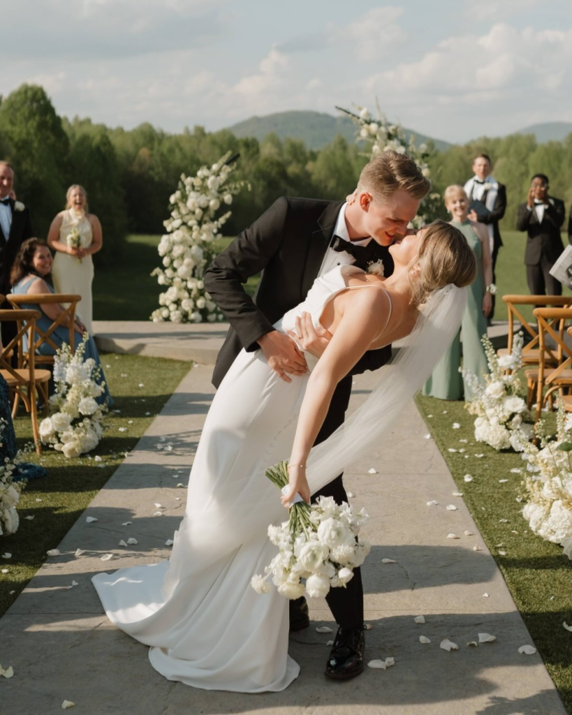 A groom dips and kisses his bride during their outdoor wedding ceremony, with guests watching in the background and floral arrangements lining the aisle.