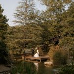 A couple in wedding attire stands on a wooden dock surrounded by lush greenery and a pond.