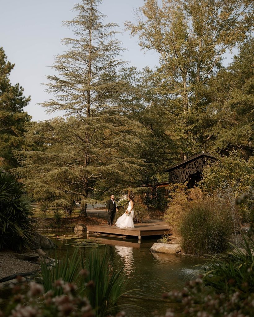 A couple in wedding attire stands on a wooden dock surrounded by lush greenery and a pond.