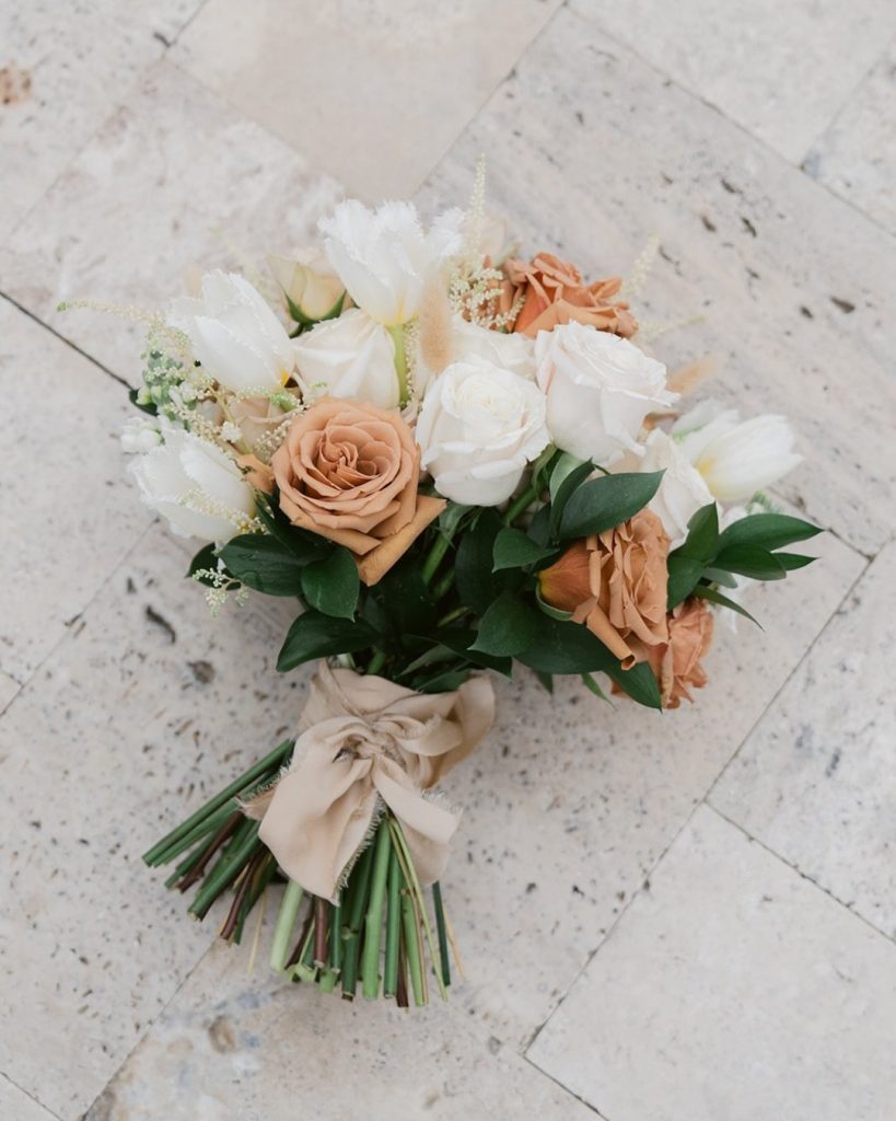 A bouquet of white and tan roses and tulips tied with a beige ribbon, resting on a stone floor.