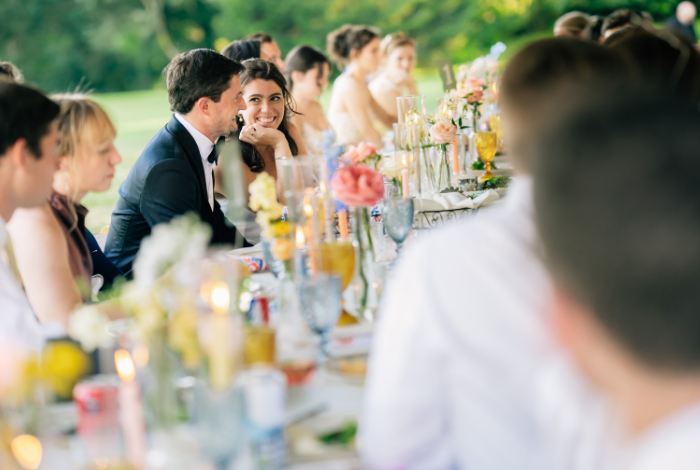 Wedding guests seated at a long reception table with colorful glassware, florals, and candles, sharing a moment during an outdoor tented dinner