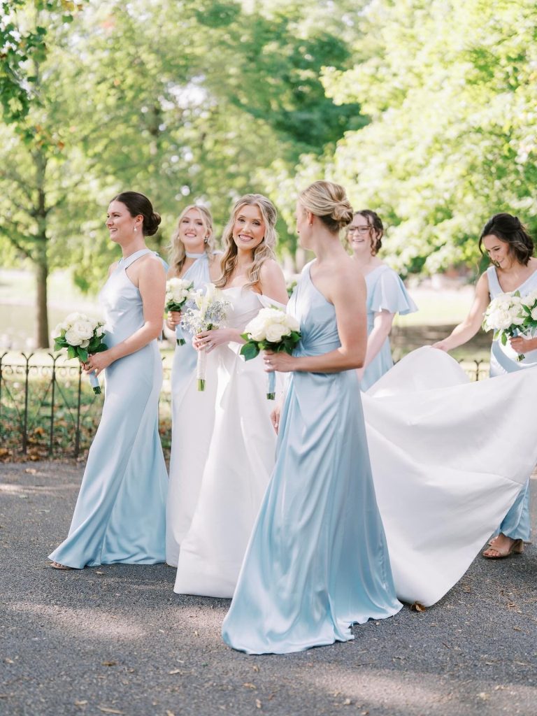 A bride smiles while walking outdoors with her bridesmaids, who are dressed in light blue and holding the bride's white gown train for her wedding planned by Hawthorne Creative Co.