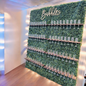 Champagne wall display in Cleveland, Ohio featuring a lush green hedge backdrop with wooden shelves lined with champagne flutes and a “Bubbles” sign above—perfect for weddings, parties, and event rentals.
