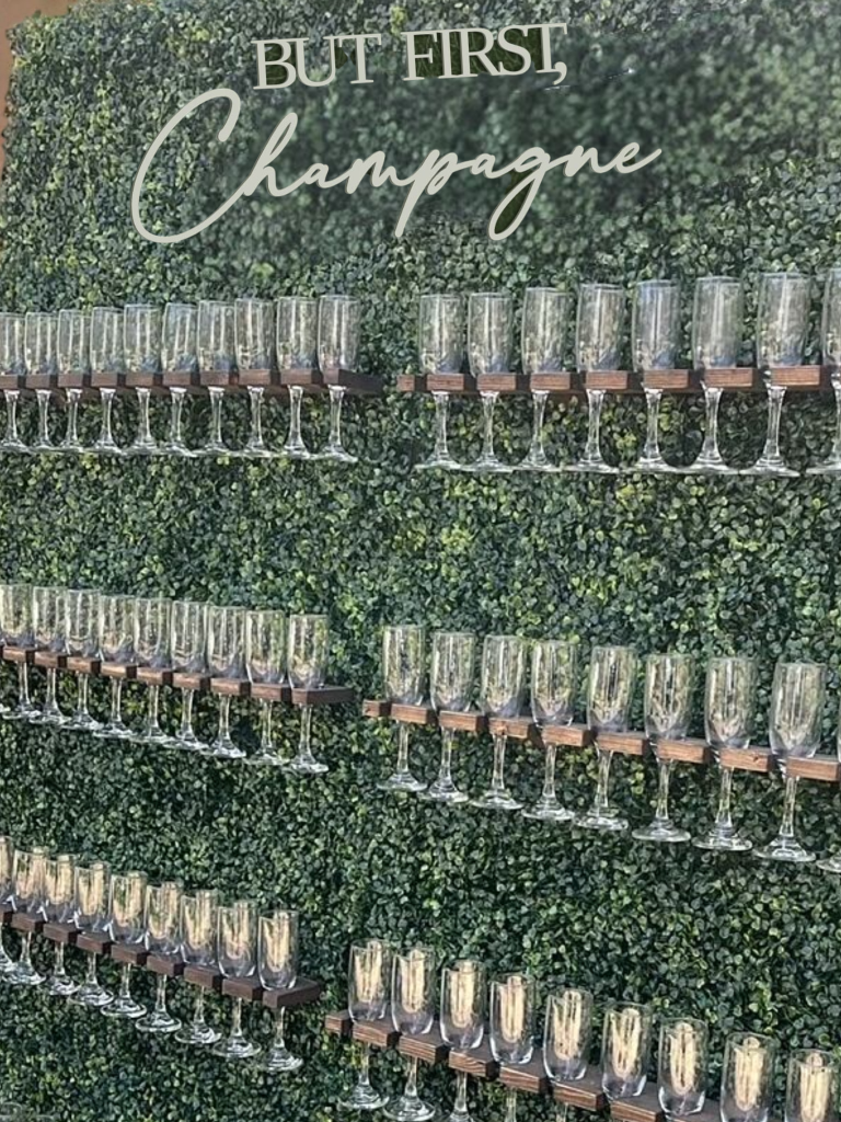 Champagne wall display in Cleveland, Ohio featuring a lush green hedge backdrop with wooden shelves lined with champagne flutes and a “Bubbles” sign above—perfect for weddings, parties, and event rentals.