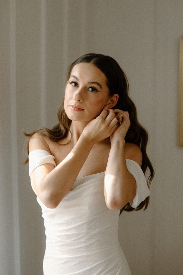 bride putting on earrings with natural wedding makeup and hollywood curls