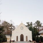 A stone chapel with arched windows and a circular window above the entrance, set against a backdrop of trees and a pale sky. A paved walkway leads to the chapel doors, with lampposts and manicured shrubs flanking the path.