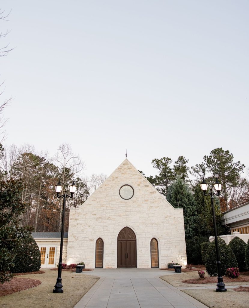A stone chapel with arched windows and a circular window above the entrance, set against a backdrop of trees and a pale sky. A paved walkway leads to the chapel doors, with lampposts and manicured shrubs flanking the path.
