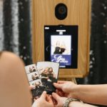 A wooden photo booth with a screen displaying two people, and hands holding out photo strips from the booth.