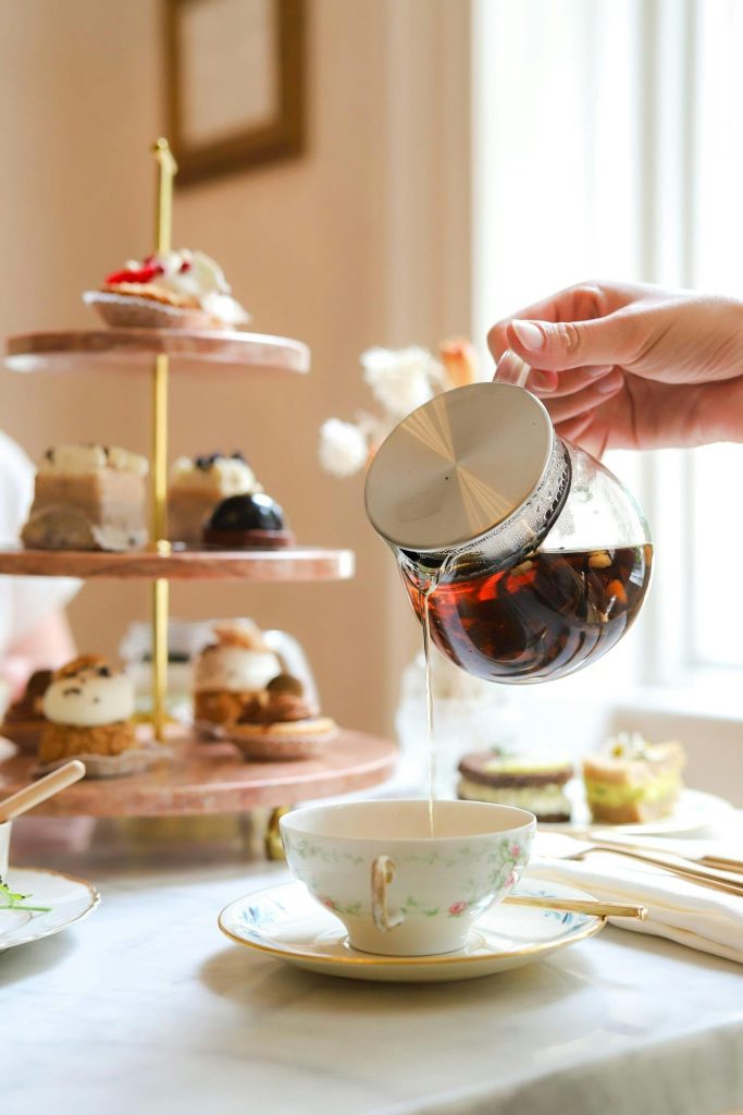tea pot with tea being poured into a tea cup with pastry's and treats in the background
