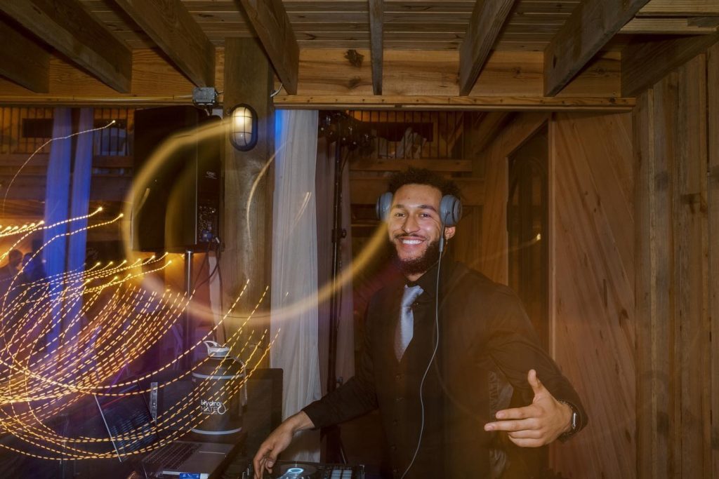 A smiling DJ with a beard and headphones poses behind a mixing console in a rustic wooden venue. Golden light trails from a long exposure swirl in the foreground.