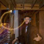 A smiling DJ with a beard and headphones poses behind a mixing console in a rustic wooden venue. Golden light trails from a long exposure swirl in the foreground.