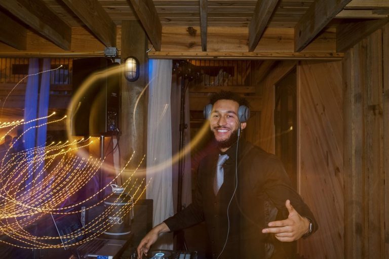 A smiling DJ with a beard and headphones poses behind a mixing console in a rustic wooden venue. Golden light trails from a long exposure swirl in the foreground.