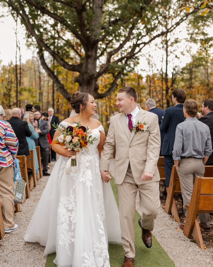 Newlywed bride and groom walking down outdoor ceremony aisle beneath a large tree at Retreat 21 in Marysville, Ohio, bride wearing floral lace wedding gown and holding orange and burgundy fall bouquet during dreamy autumn wedding celebration.
