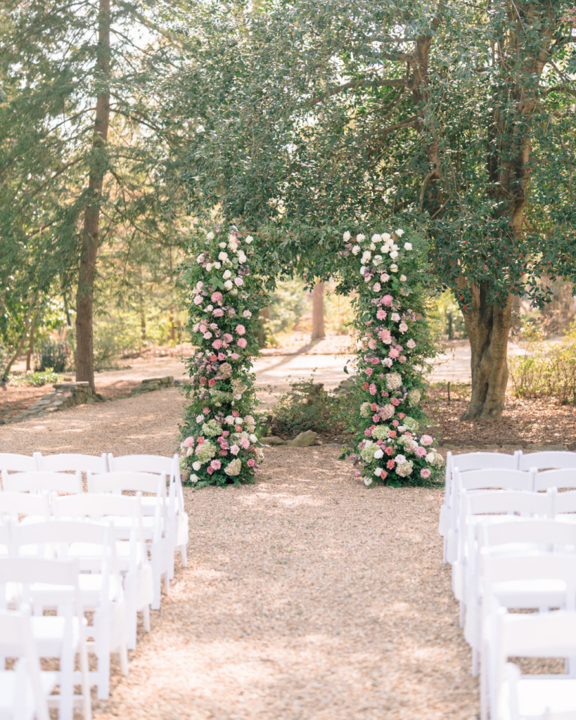 Outdoor wedding ceremony setup with white chairs and an abundant pink and cream floral arch surrounded by trees.