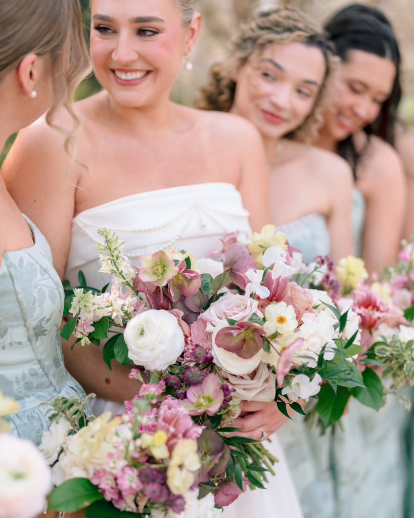 Bride smiles while holding a pastel spring bouquet alongside bridesmaids in sage floral dresses.