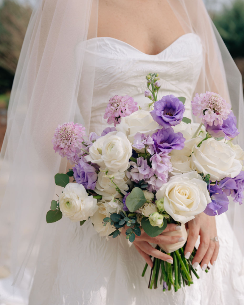 A bride holds a bouquet of white and purple flowers, including roses, ranunculus, and lisianthus, with a sheer veil draped over her shoulders.