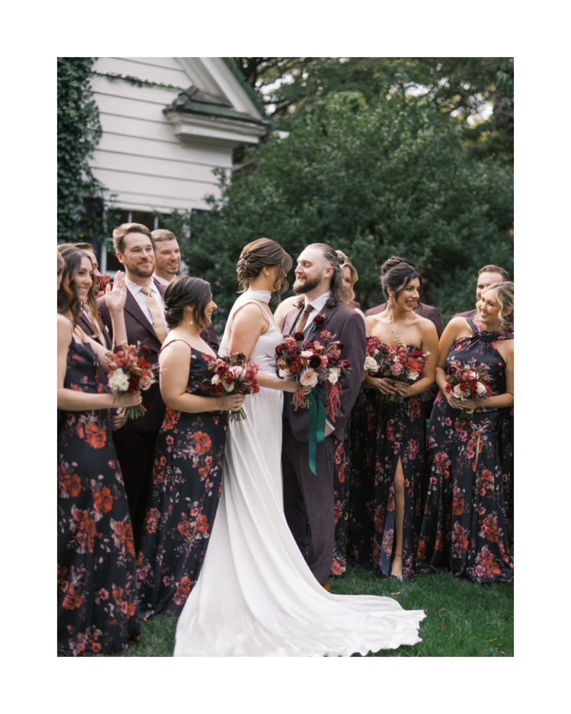 Bride and groom surrounded by bridesmaids in floral gowns and a groom in a plum suit during a romantic outdoor wedding portrait in Charlotte.
