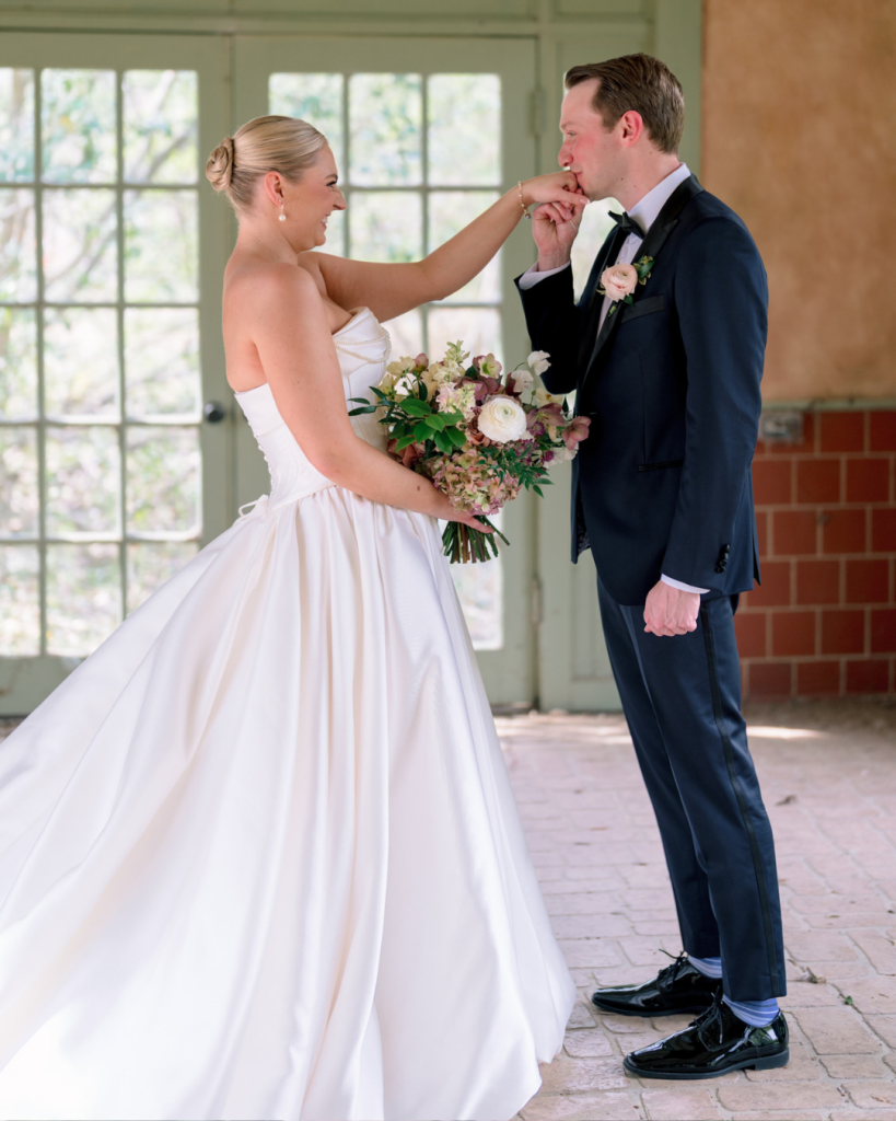 Bride smiles as groom kisses her hand during their first look at Van Landingham Estate.