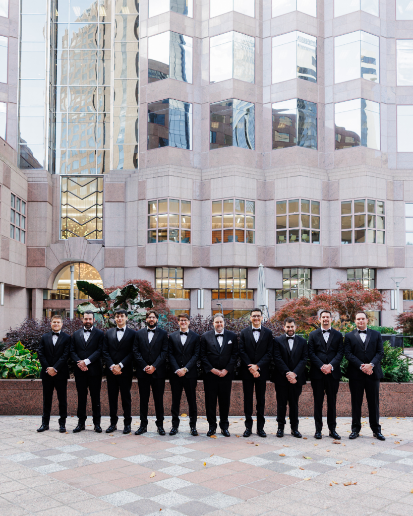 Groom and groomsmen wearing classic black tuxedos posing in Uptown Charlotte before a modern city backdrop.