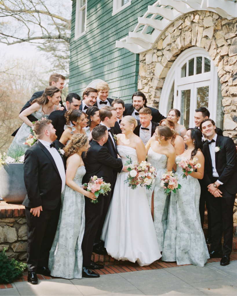 Bride and groom pose with wedding party outside Van Landingham Estate in coordinating sage floral bridesmaid dresses and black tuxedos.