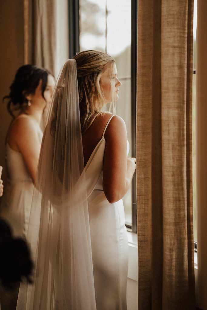 Bride in satin wedding gown looking out window with bridesmaid during getting ready moments at Delaware wedding venue