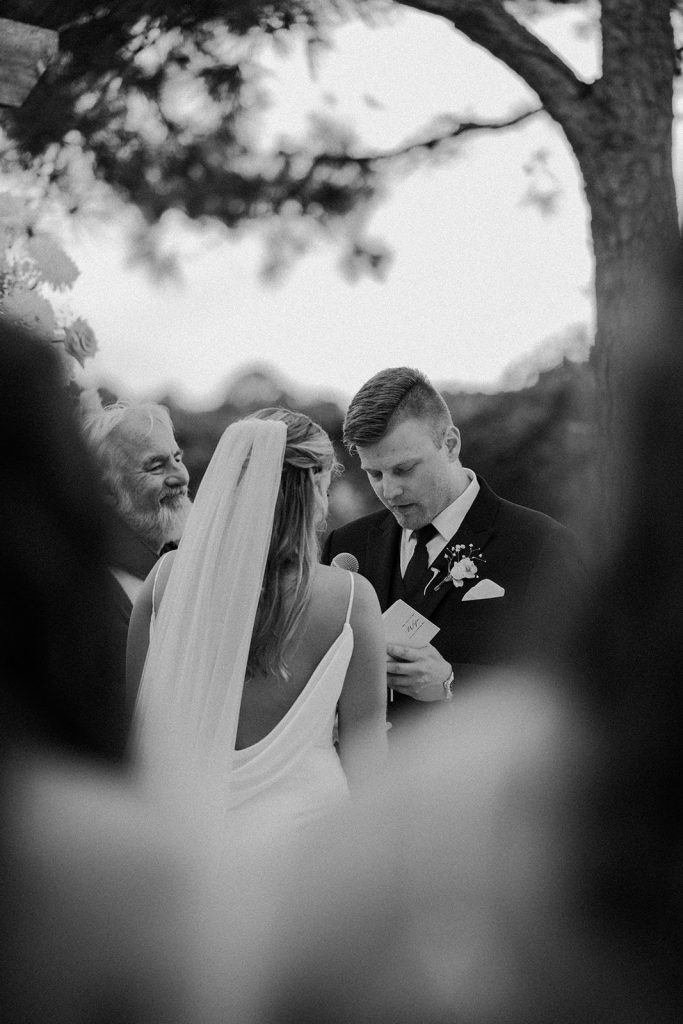 Black-and-white photo of groom reading handwritten vows to bride during an outdoor waterfront wedding ceremony at Bayside Resort Golf Club in Selbyville, Delaware.