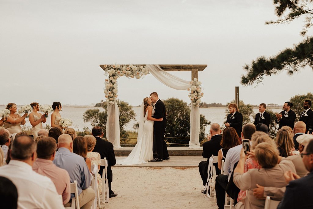 Molly and Curtis sharing their first kiss beneath a draped floral ceremony arch during their waterfront wedding ceremony at Bayside Resort Golf Club in Selbyville, Delaware.