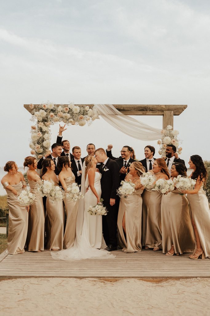 Wedding party cheering around Molly and Curtis during their beachside wedding ceremony at Bayside Resort Golf Club in Selbyville, Delaware, with neutral bridesmaid dresses, black tuxedos, and a floral ceremony arch overlooking the bay.