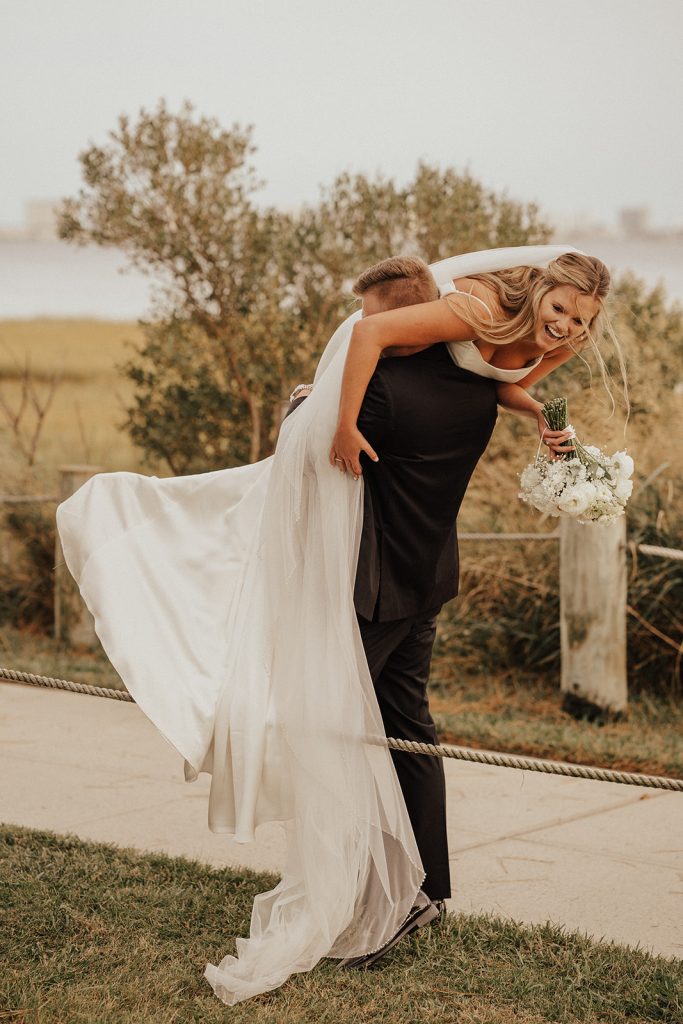 Groom carrying bride on his back along a coastal pathway at Bayside Resort Golf Club in Selbyville, Delaware, during a playful sunset wedding portrait session.