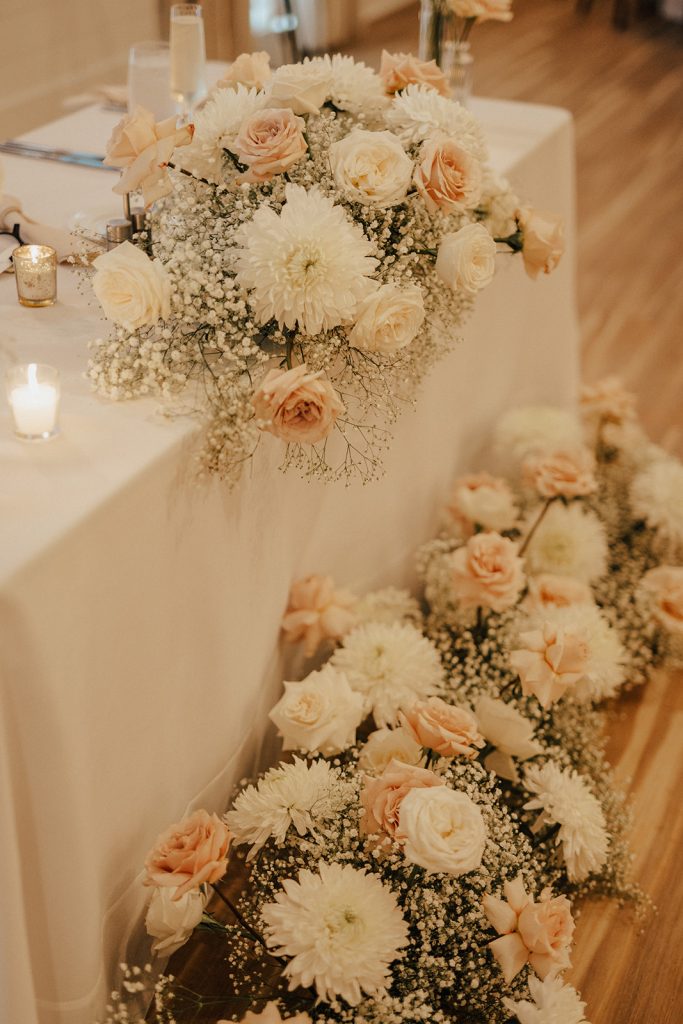Low floral arrangement with white chrysanthemums, blush roses, and baby’s breath decorating the sweetheart table at a Bayside Resort Golf Club wedding reception in Selbyville, Delaware.