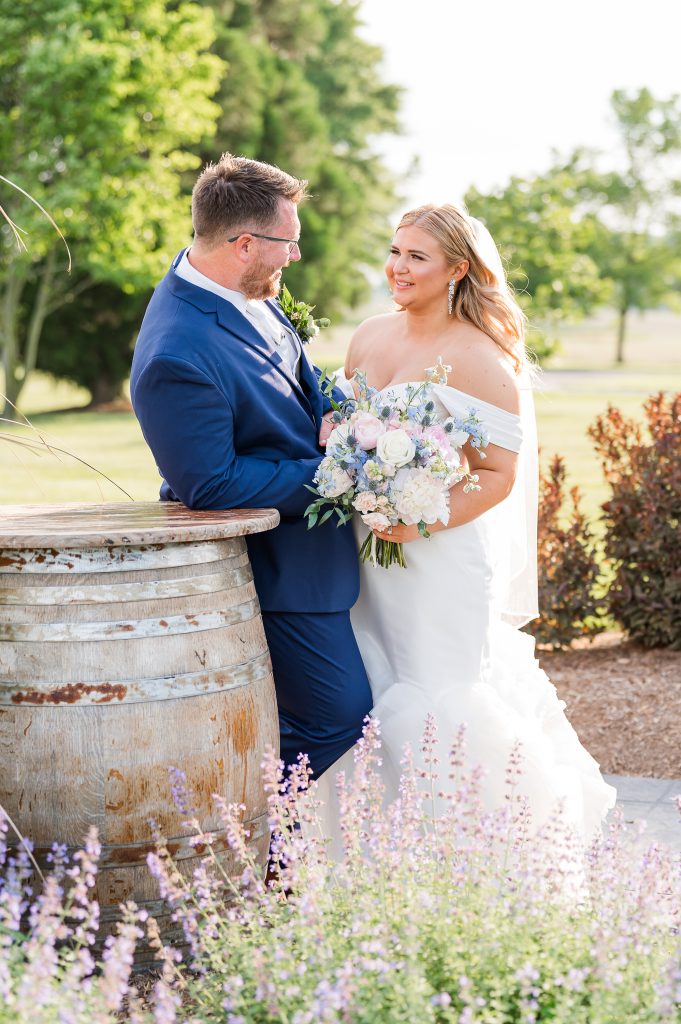 Bride and groom leaning against weathered barrels surrounded by lavender and garden flowers during sunset portraits at King Cole Farm.