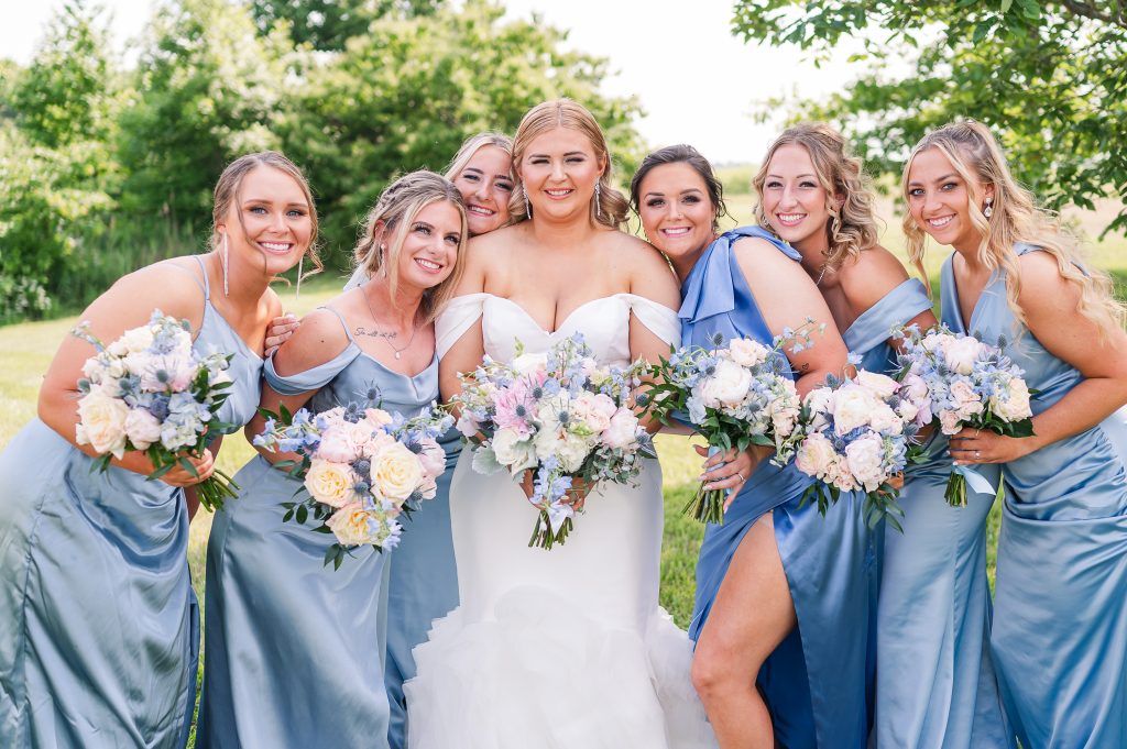 Bride posing and laughing with bridesmaids in matching satin blue dresses during wedding portraits at King Cole Farm.