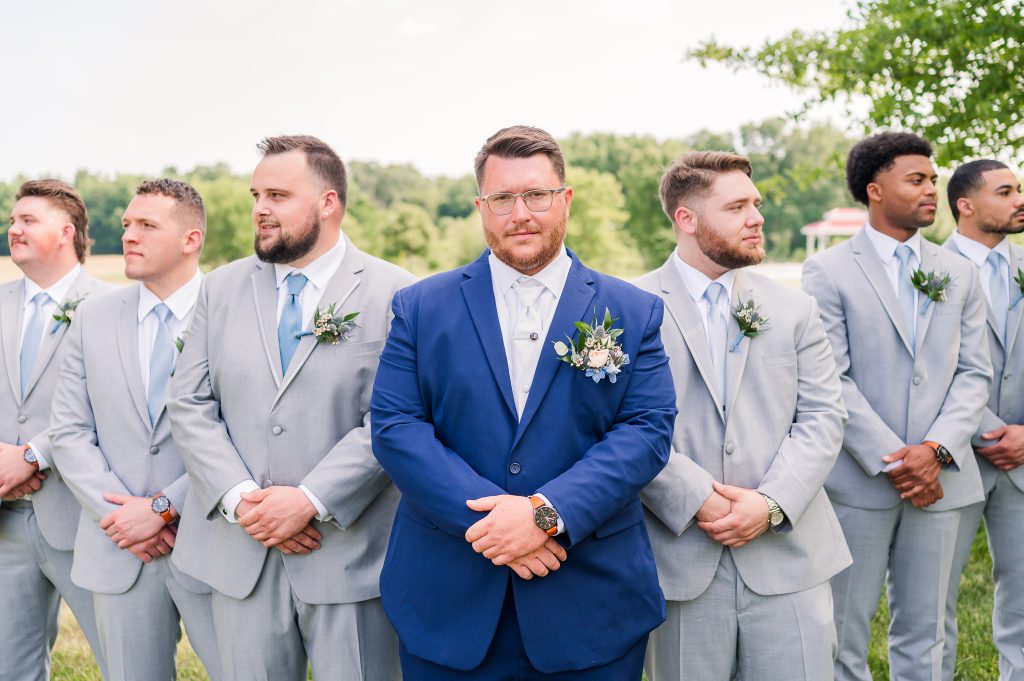Groom in a navy suit standing with gray-suited groomsmen during wedding party portraits at King Cole Farm in Delaware.