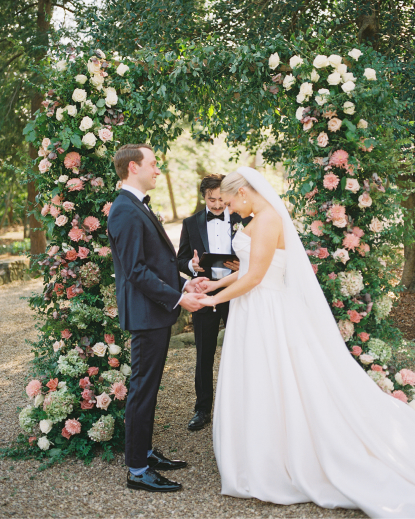 Bride and groom exchange vows beneath a lush floral ceremony arch during their outdoor garden wedding ceremony.