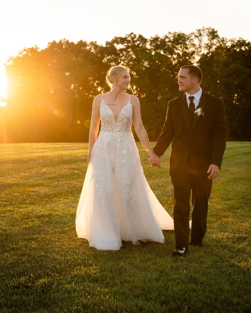 Bride and groom walking hand in hand through a sunlit field at golden hour during their October wedding in Elkton, Maryland.