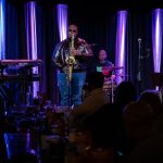 Anthony Saxson performing saxophone alongside a live band under purple stage lighting.