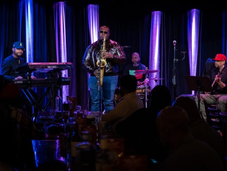 Anthony Saxson performing saxophone alongside a live band under purple stage lighting.