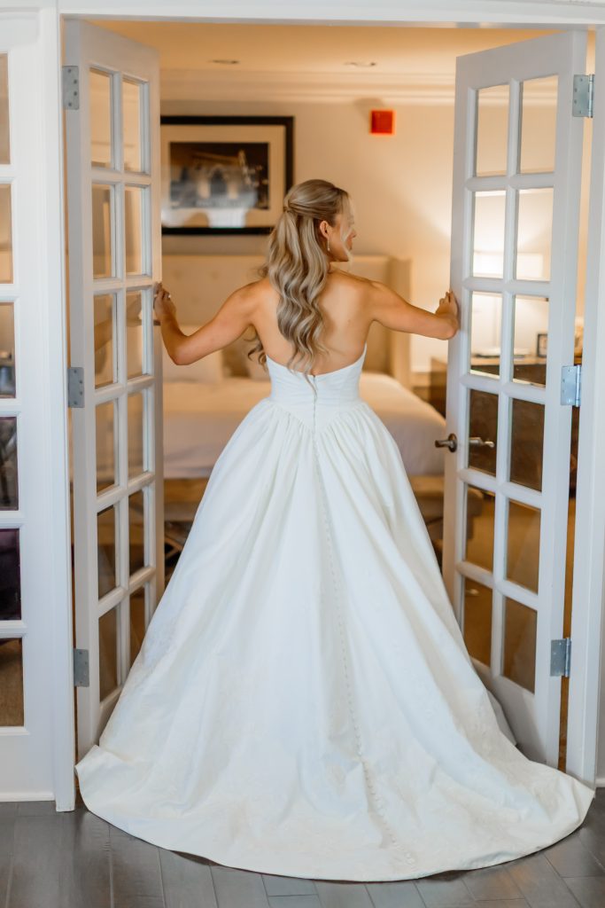 Behind-the-shot of the bride in her gown altered by Alterations by Debbie, standing between French doors and looking into her bridal suite.