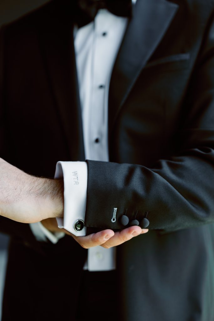 Close-up of a groom adjusting black cufflinks on a classic black tuxedo jacket.