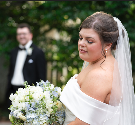 A bride in a white off-the-shoulder wedding dress holds a bouquet of white and blue flowers. In the blurred background, a person in a tuxedo stands.