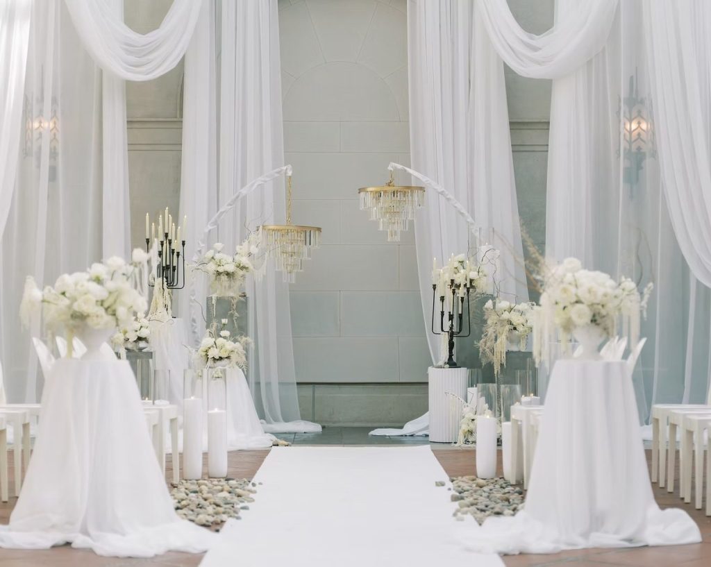 Elegant all-white wedding ceremony setup featuring flowing floor-to-ceiling drapery, modern curved arches with crystal chandeliers, lush white floral arrangements, black candelabras, candle-lined aisle, and a white aisle runner creating a luxurious, romantic ceremony backdrop.