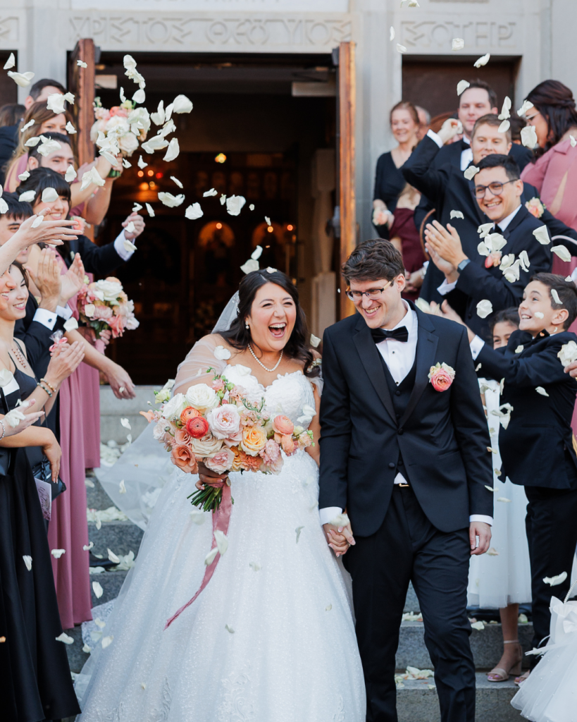 Bride and groom exit Holy Trinity Greek Orthodox Cathedral in Charlotte, North Carolina while guests toss white flower petals during a joyful wedding celebration photographed by ParksPictures for Wed Society Charlotte.
