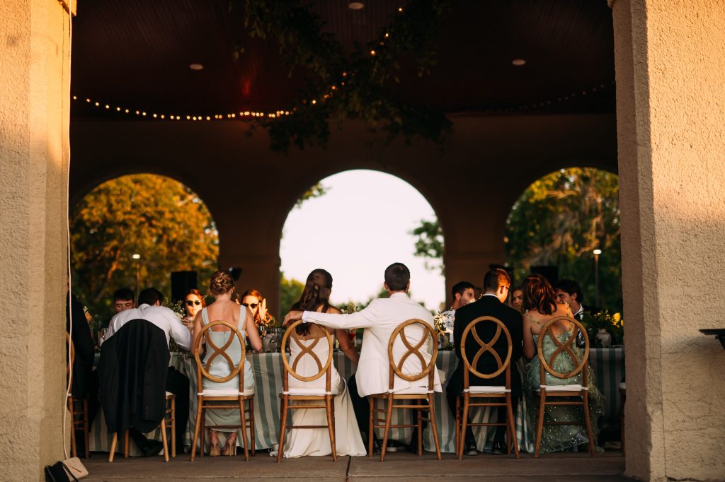 Bride and groom sitting at head table at World's Fair Pavilion with string lights on ceiling and wooden chairs from Exclusive Events.