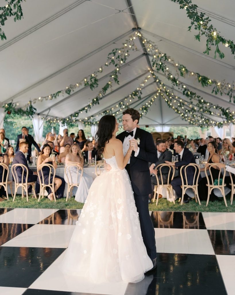 Bride and groom dancing on a black and white checkered floor under a tent with string lights.