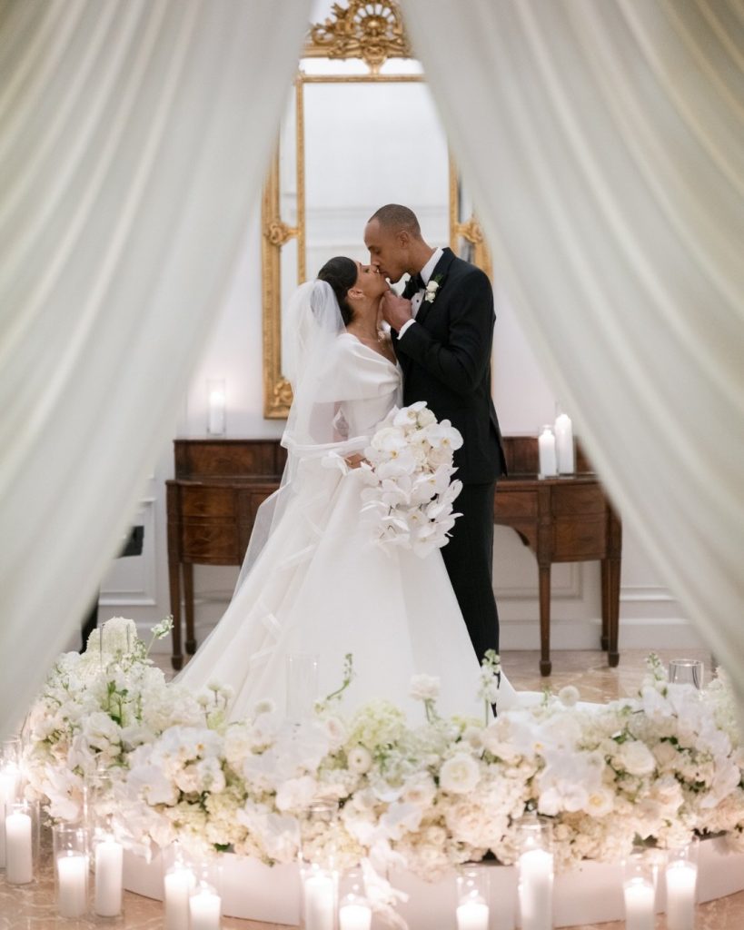 A bride in a white gown and veil kisses the groom in a black tuxedo, surrounded by abundant white floral arrangements and lit candles.