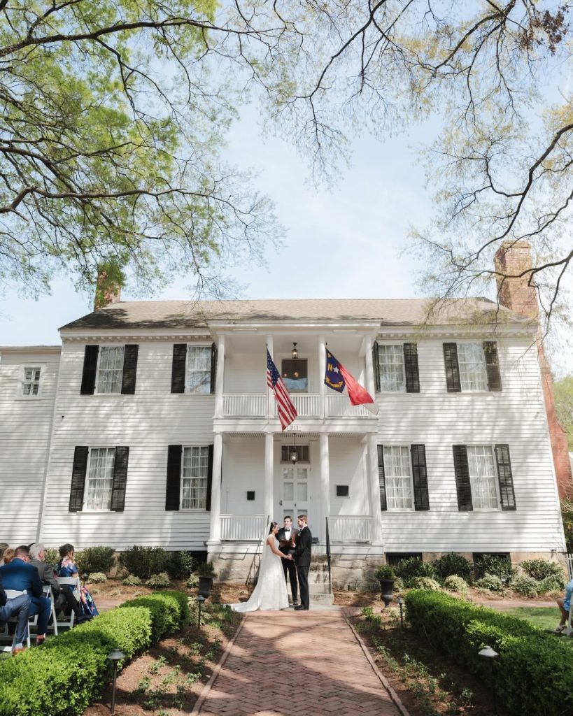 Wedding ceremony taking place on the steps of a two-story white house with black shutters and flags.