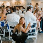 Content Creator at a wedding reception smiling toward the camera while guests sit and mingle in the background inside a bright event space.