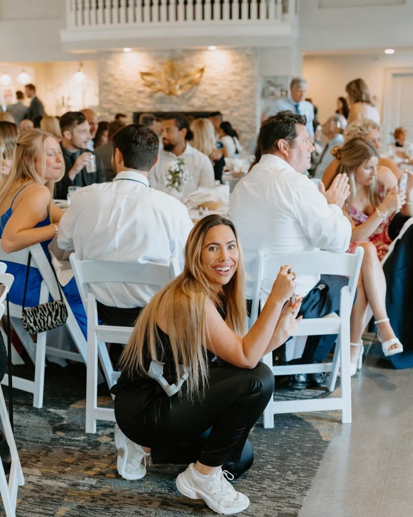 Content Creator at a wedding reception smiling toward the camera while guests sit and mingle in the background inside a bright event space.