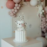 A two-tier white cake with a fluted bottom layer, pink monogram, and delicate dried daisy floral accents.