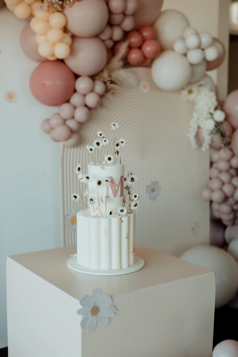 A two-tier white cake with a fluted bottom layer, pink monogram, and delicate dried daisy floral accents.